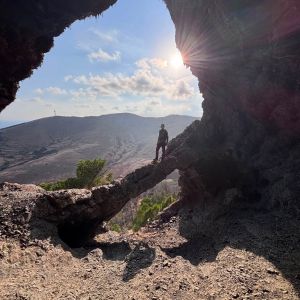 A man in a hat stands alone on a rock looking out of a cave
